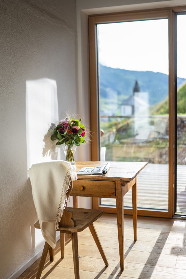 Kleiner Holztisch mit Stuhl, Blumenstrauß und Blick durch die Terrassentür auf die Berge