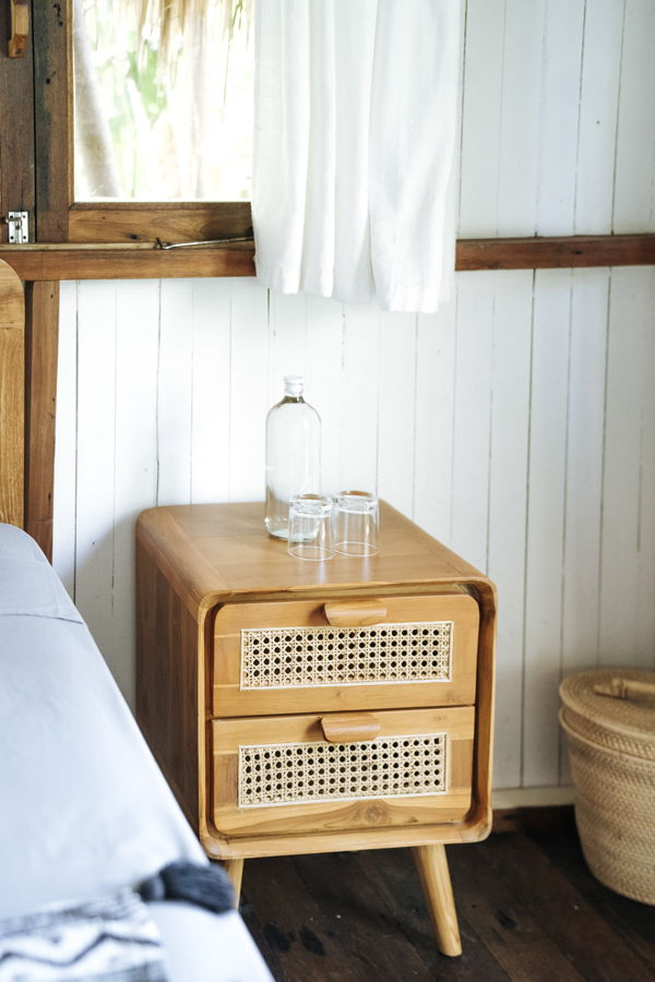 Holz-Nachttisch mit zwei Schubladen neben einem Bett, darauf eine Flasche und zwei Gläser