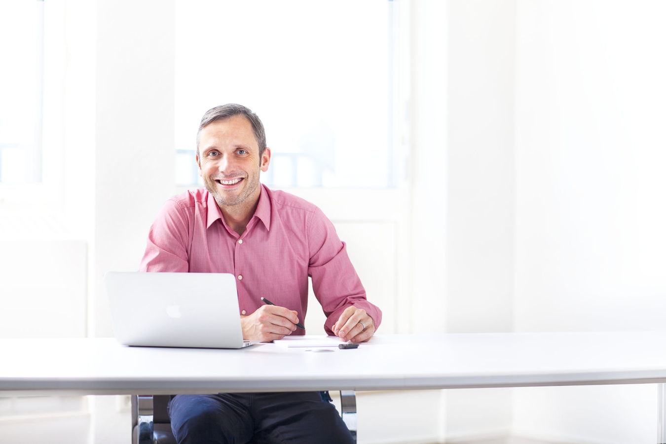 Mann in rosa Hemd sitzt am Schreibtisch mit Laptop und Stift