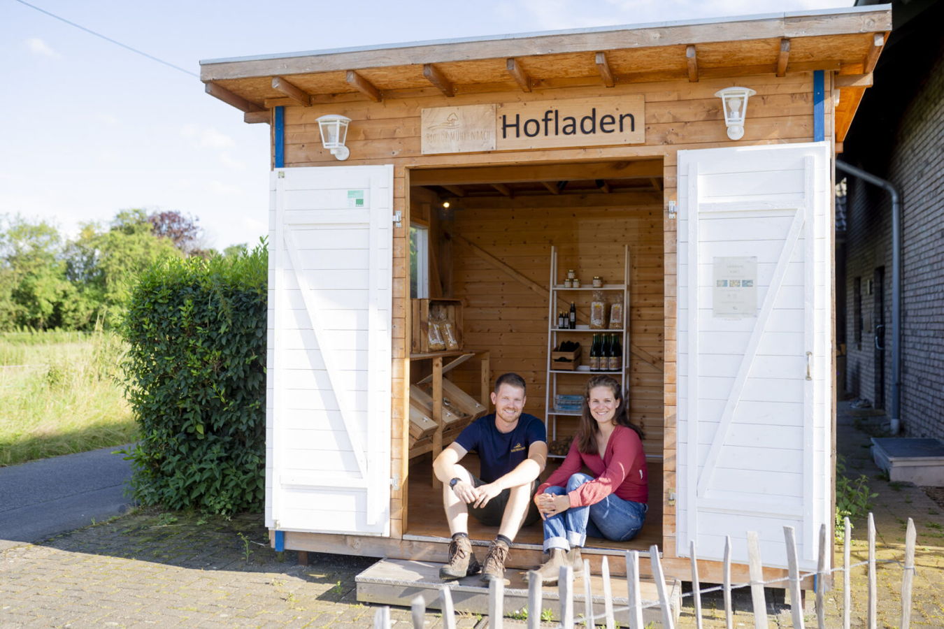 Zwei Personen sitzen vor einem Holz-Hofladen mit geöffneten Türen
