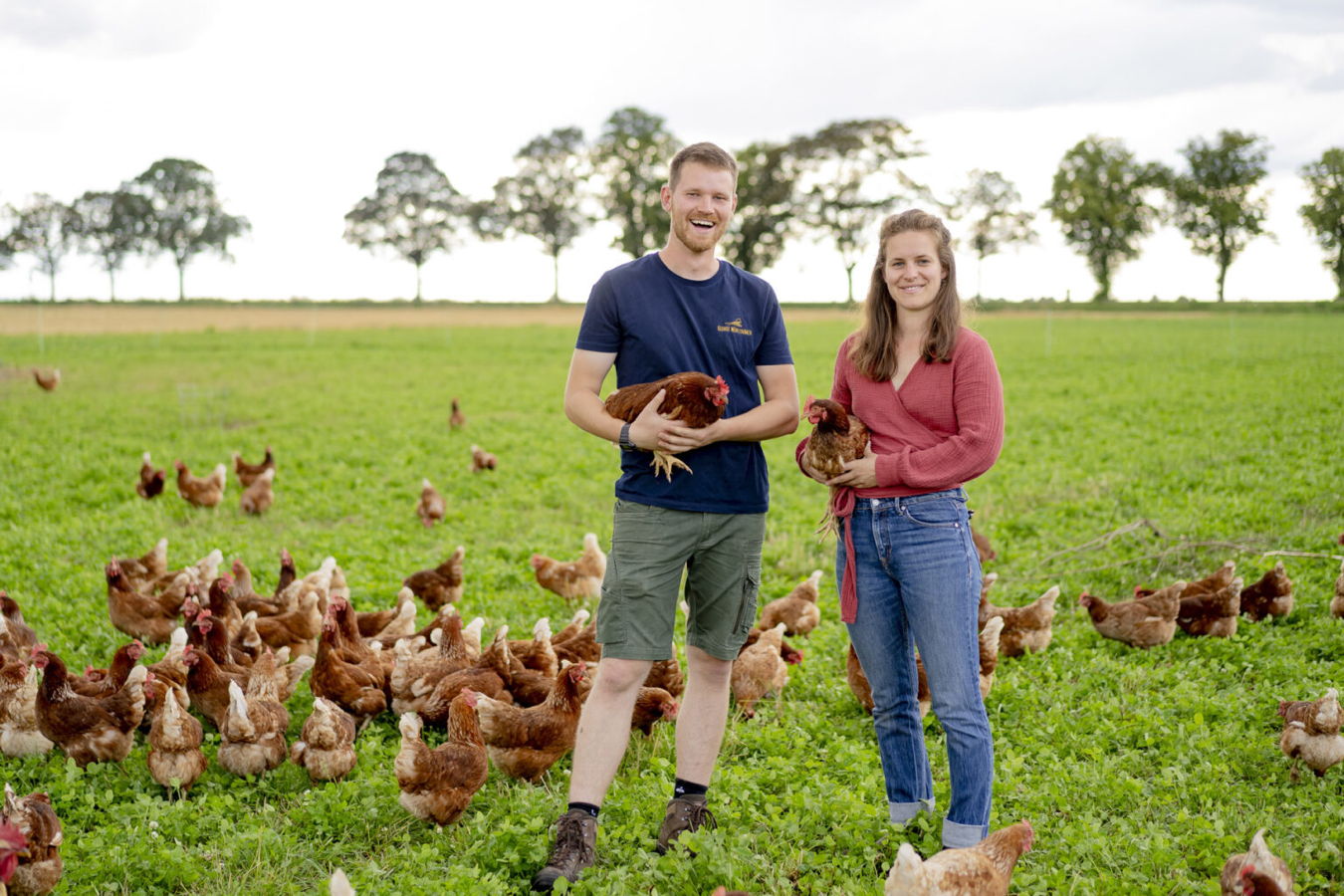 Ein Mann und eine Frau stehen auf einer Wiese zwischen vielen Hühnern und halten jeweils ein Huhn im Arm