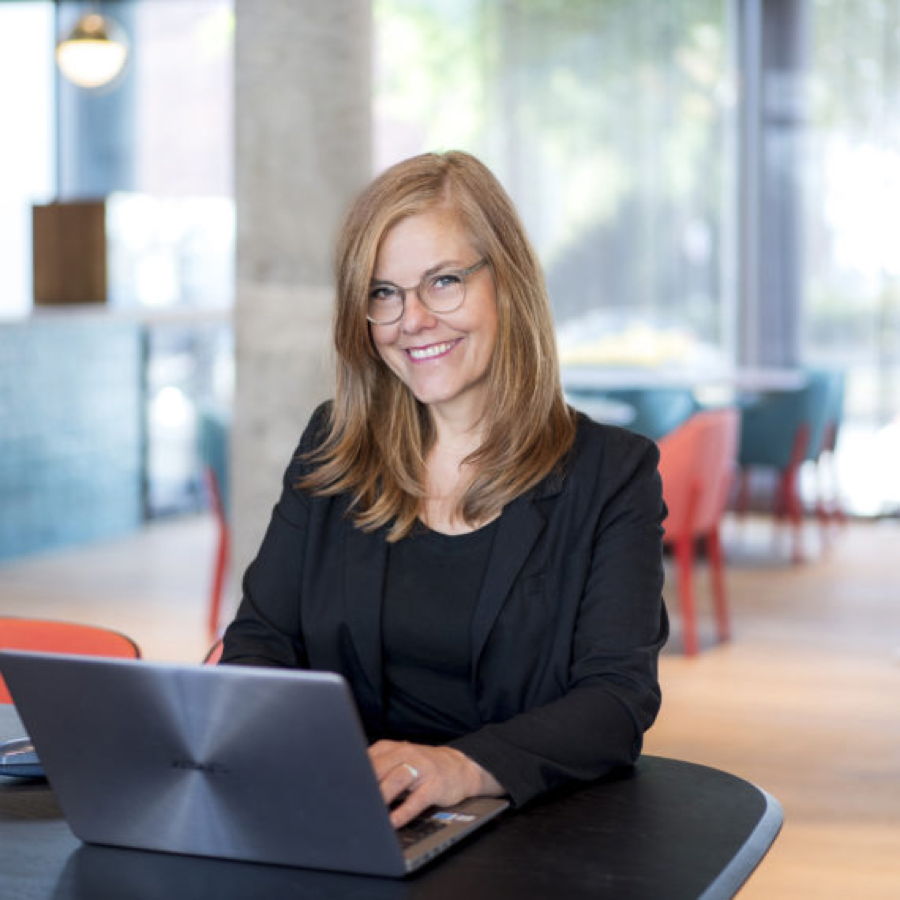 Eine lächelnde Frau mit Brille arbeitet an einem Laptop in einem modernen Café