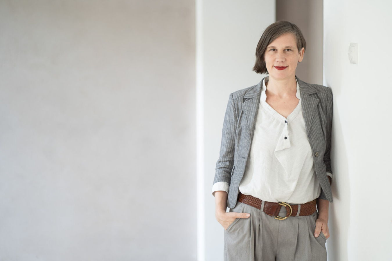 Frau in grauem Blazer und weißer Bluse lehnt an einer weißen Wand
