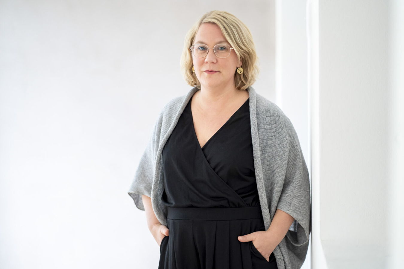 Porträt einer Frau mit Brille in schwarzem Kleid und grauem Schal vor hellem Hintergrund