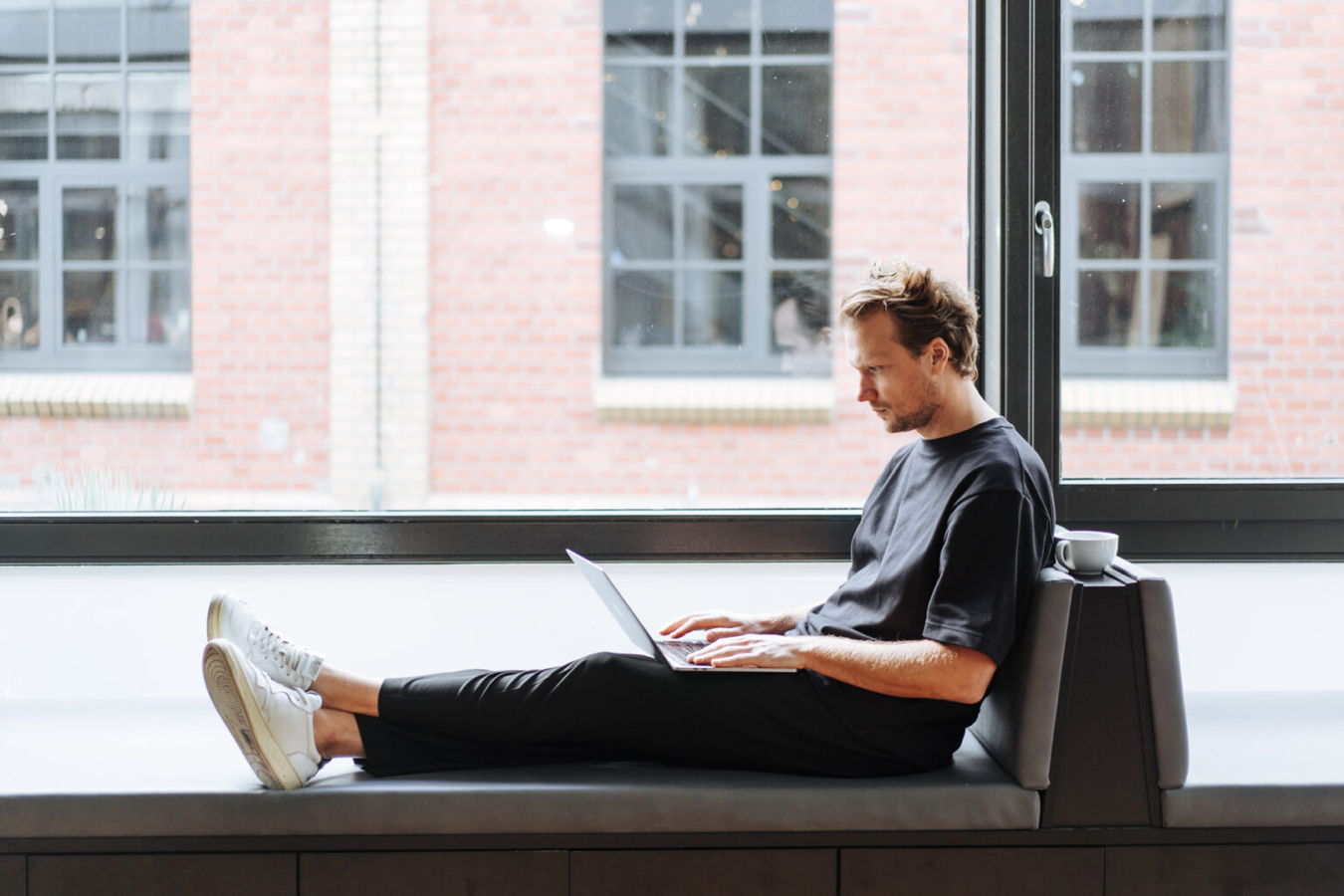 Mann sitzt mit Laptop auf einer Fensterbank und arbeitet