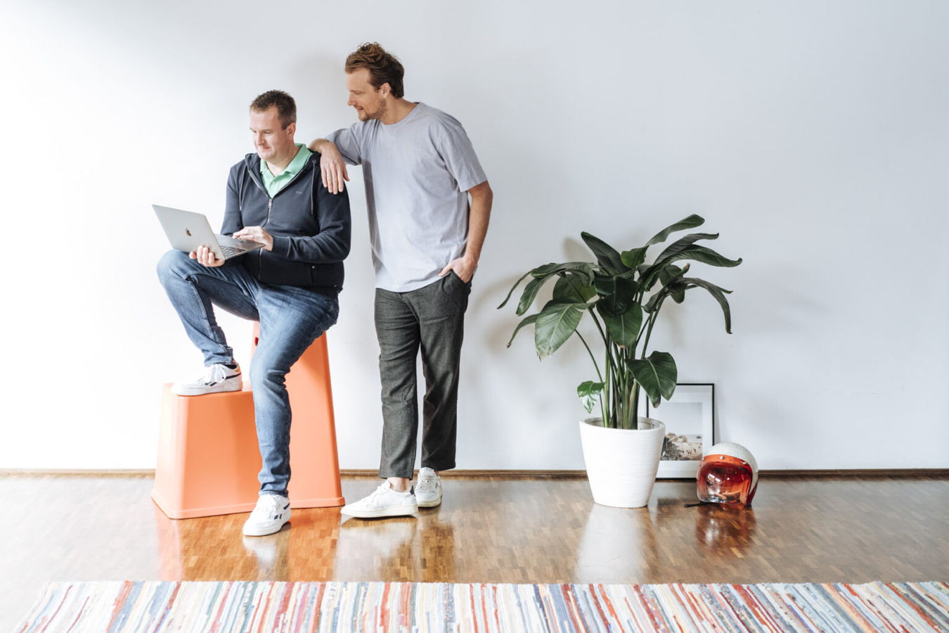 Zwei Männer in einem hellen Raum; einer sitzt mit Laptop auf einem Hocker, der andere lehnt daneben