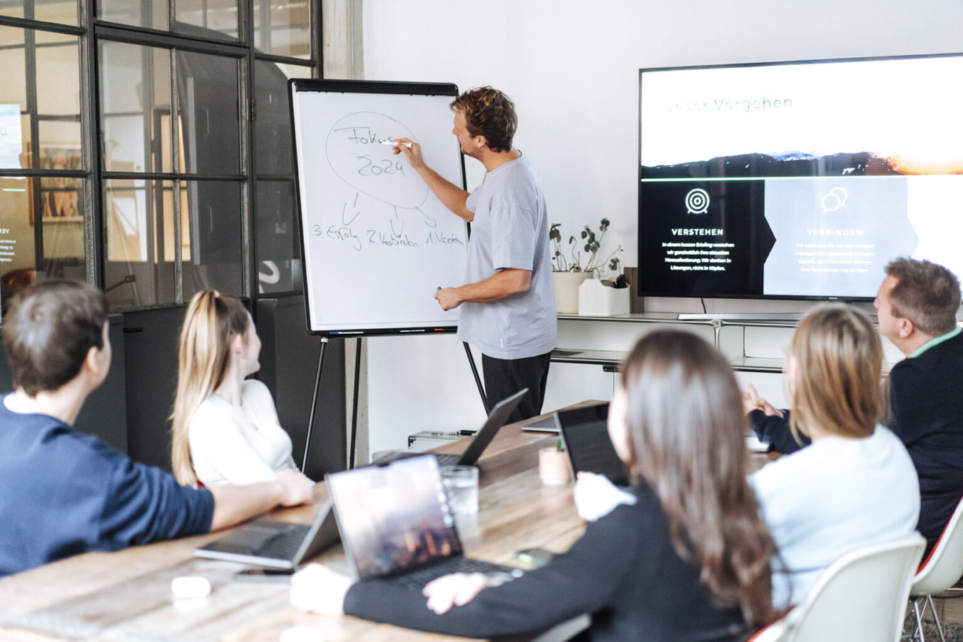 Mann präsentiert vor einem Flipchart in einer Besprechung mit mehreren Zuhörern am Tisch