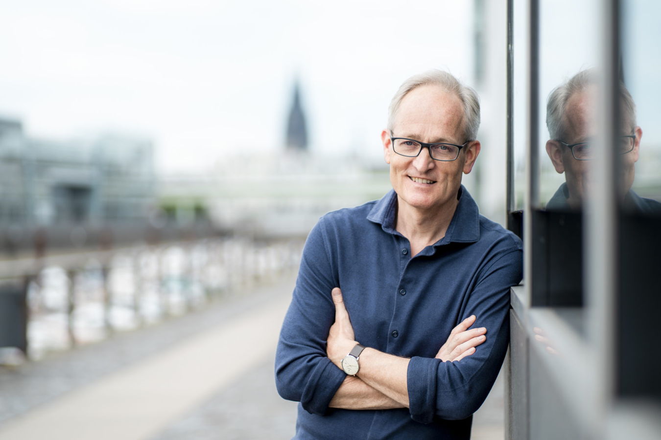 Porträt eines lächelnden Mannes mit Brille, der an einer Glaswand lehnt