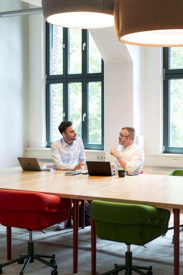 Zwei Männer besprechen sich an einem Konferenztisch mit Laptops in einem hellen Besprechungsraum