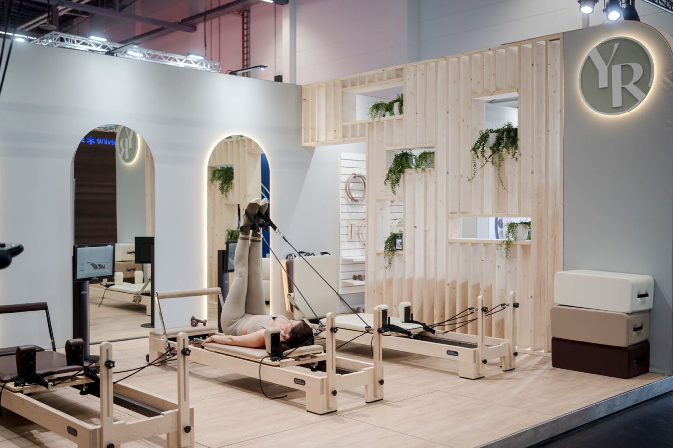 Person trainiert auf einem Pilates-Reformer in einem hellen Messestand mit Holzverkleidung