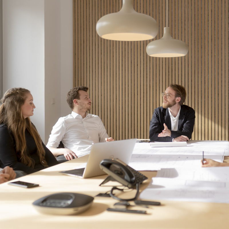 Drei Personen sitzen in einer Besprechung an einem Tisch mit Laptop und Telefon