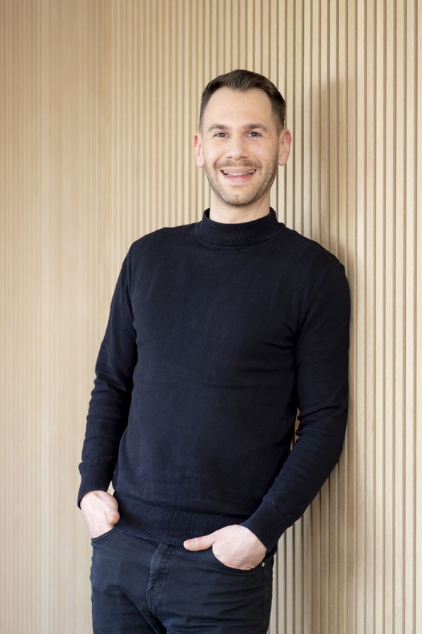 Mann in schwarzem Pullover lehnt lächelnd an einer Holzvertäfelung