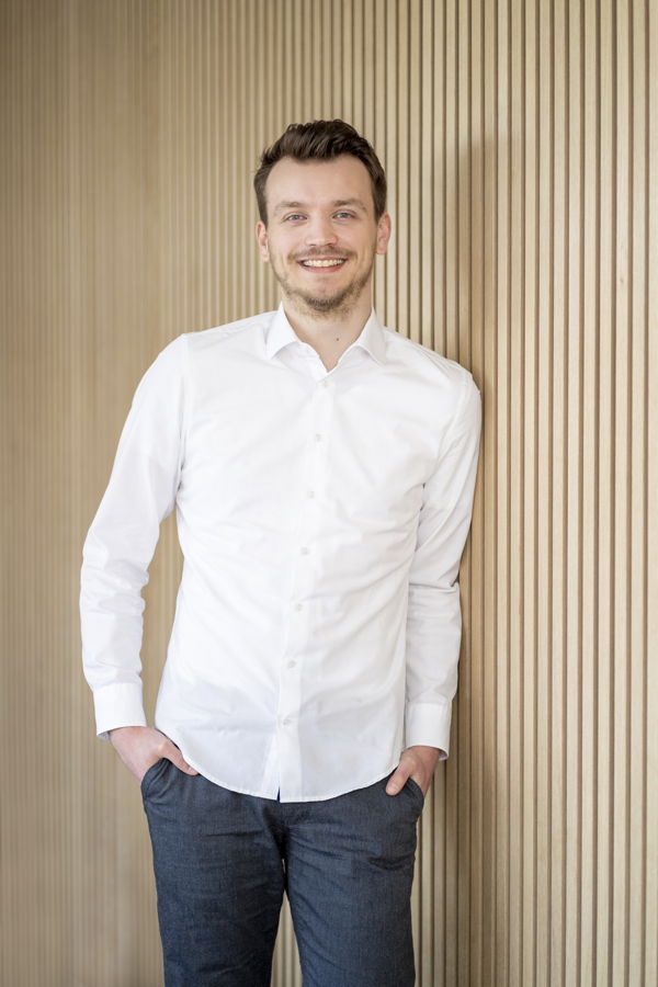 Mann in weißem Hemd und Jeans lehnt lächelnd an einer Holzlamellenwand