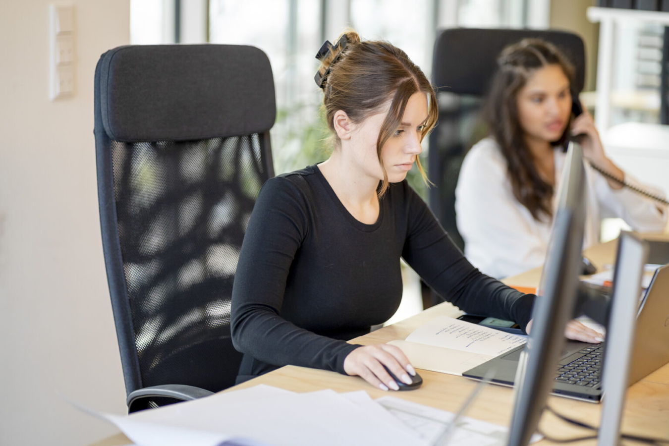 Zwei Frauen arbeiten im Büro am Computer, eine telefoniert im Hintergrund