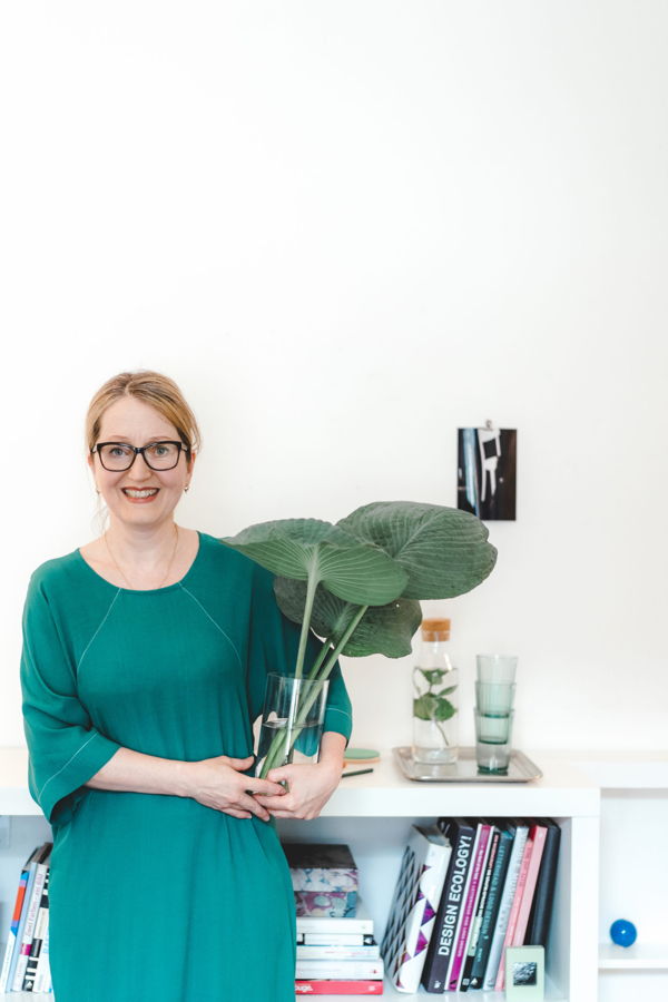 Frau in grünem Kleid hält eine Vase mit großen grünen Blättern vor einem Regal