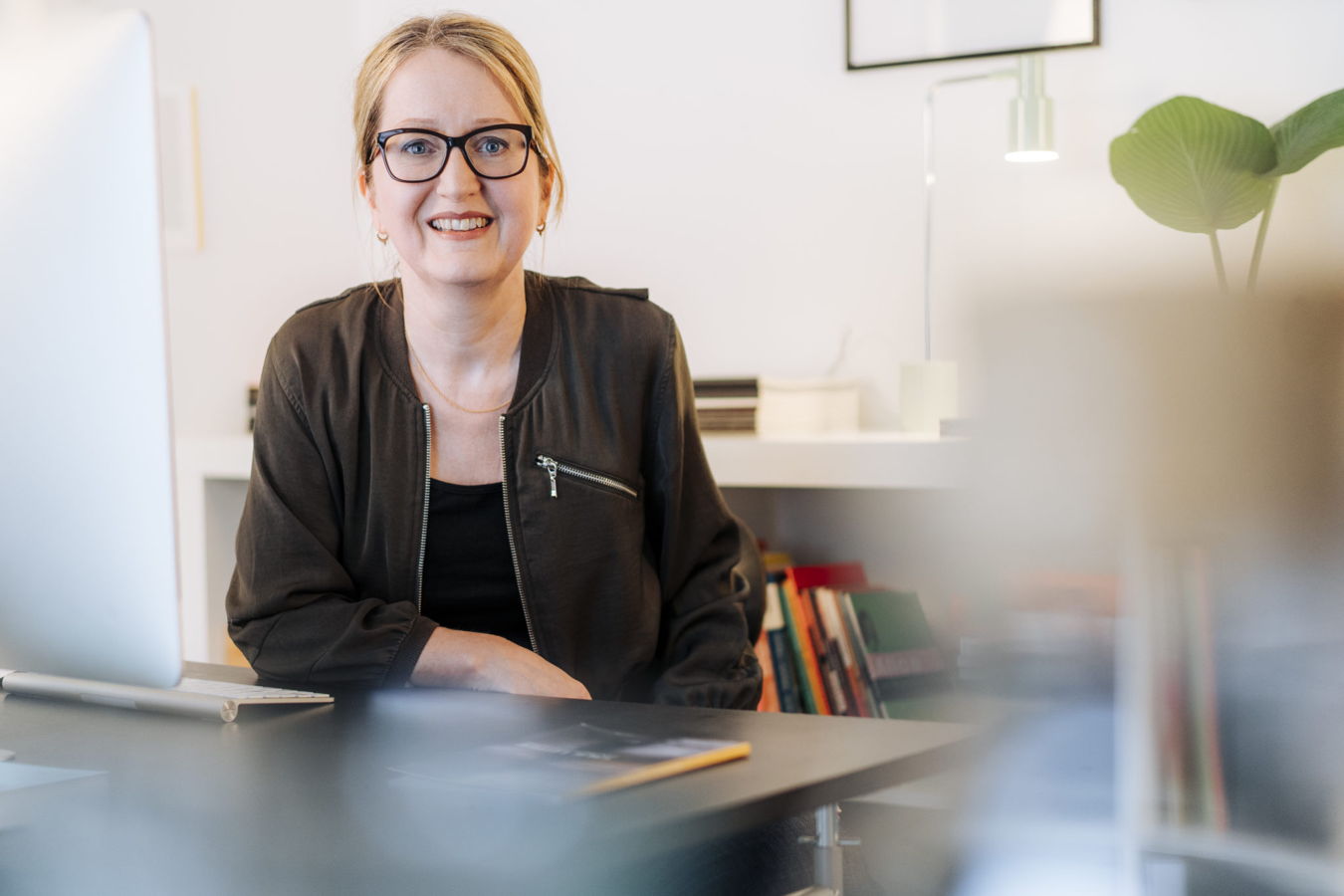 Frau mit Brille sitzt lächelnd an einem Schreibtisch im Büro