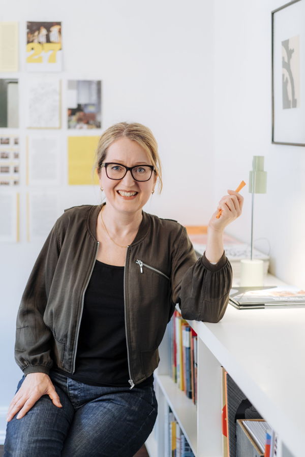 Frau mit Brille sitzt in einem hellen Arbeitszimmer und hält einen orangefarbenen Stift