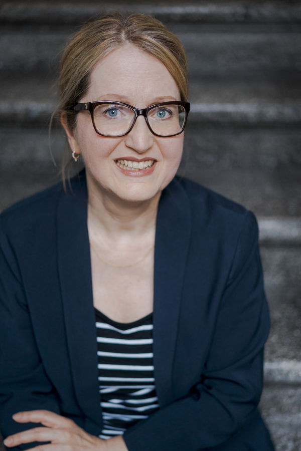 Porträt einer lächelnden Frau mit Brille und dunkelblauem Blazer vor einem dunklen Hintergrund