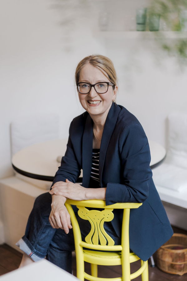 Porträt einer lächelnden Frau mit Brille, die auf einem gelben Stuhl sitzt