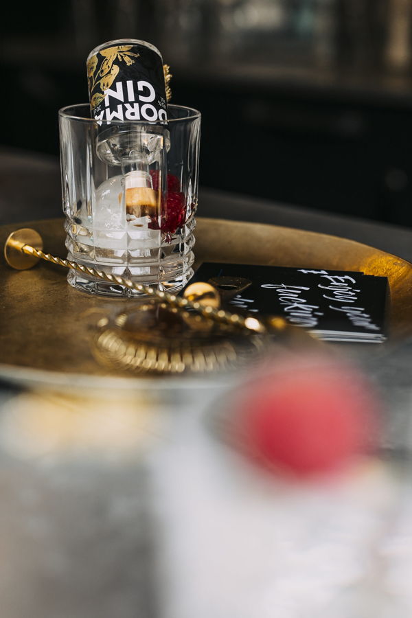 Ein Glas mit Eis und einer umgedrehten Gin-Dose auf einem Tablett, daneben ein goldener Rührlöffel und eine Karte