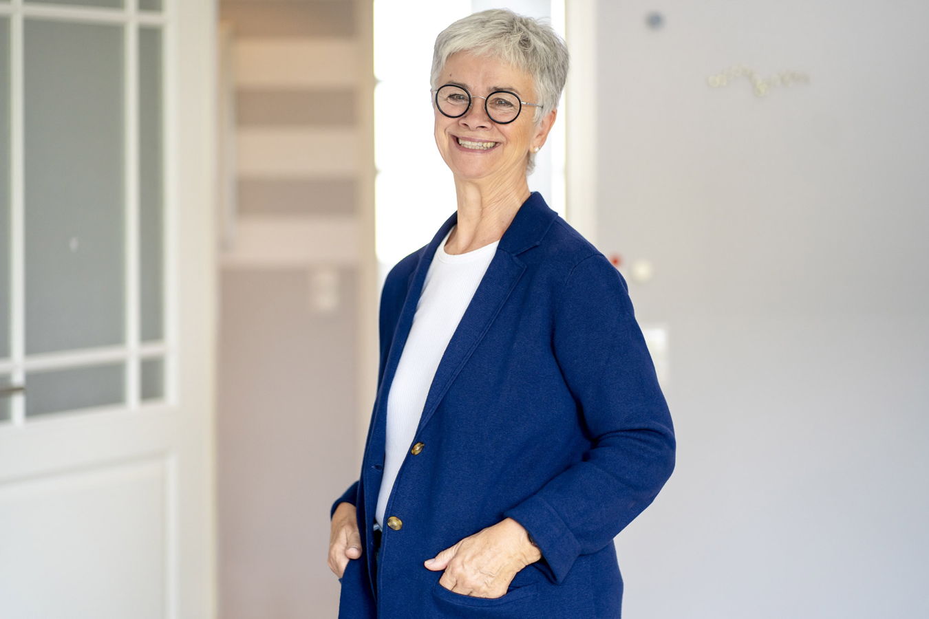 Lächelnde ältere Frau mit kurzen grauen Haaren und Brille in blauem Blazer