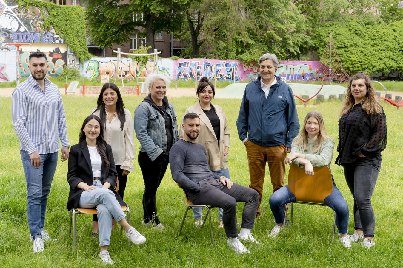 Gruppe von acht Personen steht und sitzt auf einer Wiese vor einer bunten Graffiti-Wand im Park