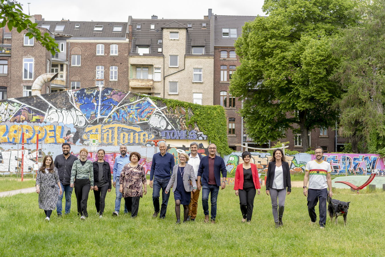 Gruppe von zwölf Personen geht über eine Wiese vor einer bunten Graffitiwand und Wohnhäusern
