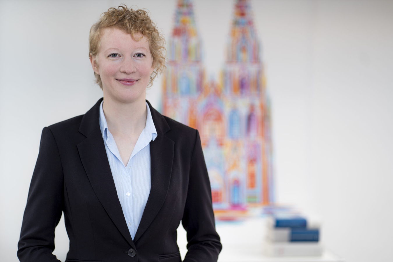Porträt einer Frau im schwarzen Blazer vor einem farbigen, unscharfen Hintergrundbild