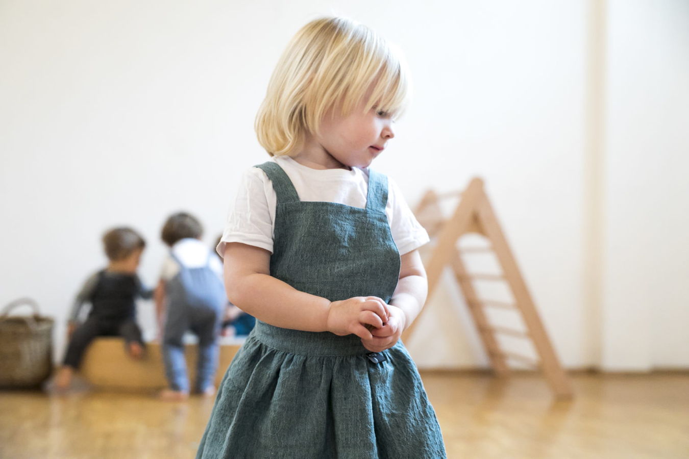 Kleines blondes Kind im grünen Kleid steht in einem hellen Raum; im Hintergrund spielen weitere Kinder