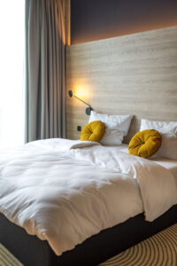 Gemachtes Bett mit weißen Laken, gelben Kissen und Wandlampe am Fenster