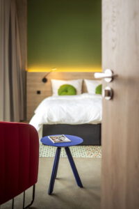 Hotelzimmer mit Bett, blauem Beistelltisch und rotem Sessel