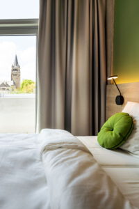 Hotelzimmer mit Bett, grünem Kissen und Blick auf einen Kirchturm durchs Fenster