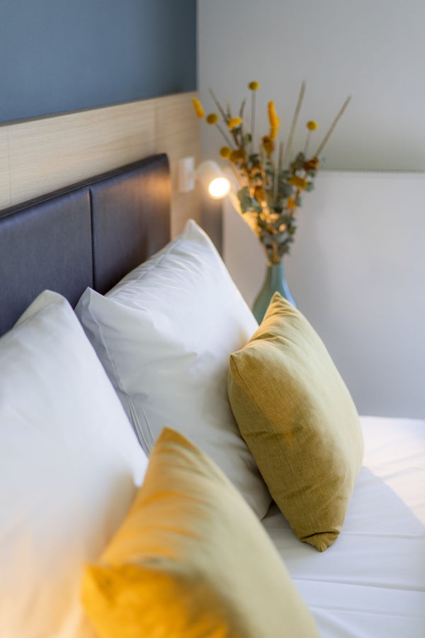 Gemachtes Bett mit weißen und gelben Kissen neben einer Vase mit Trockenblumen