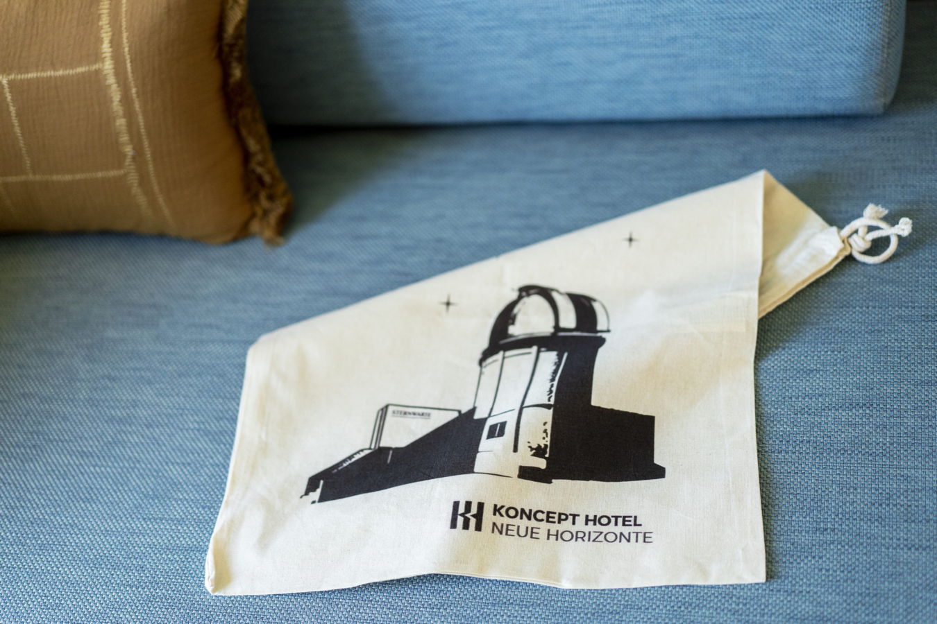 Stoffbeutel mit dem Aufdruck „Koncept Hotel Neue Horizonte“ auf einem blauen Sofa