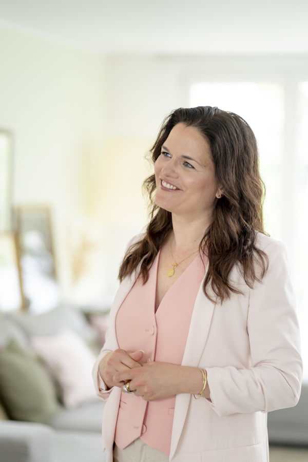 Eine lächelnde Frau in einem hellen Raum, mit rosa Bluse und weißem Blazer