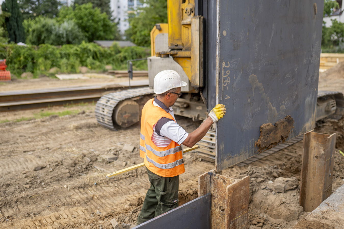 Bauarbeiter in Warnweste und Helm arbeitet an einer großen Stahlplatte neben einer Baumaschine auf einer Baustelle