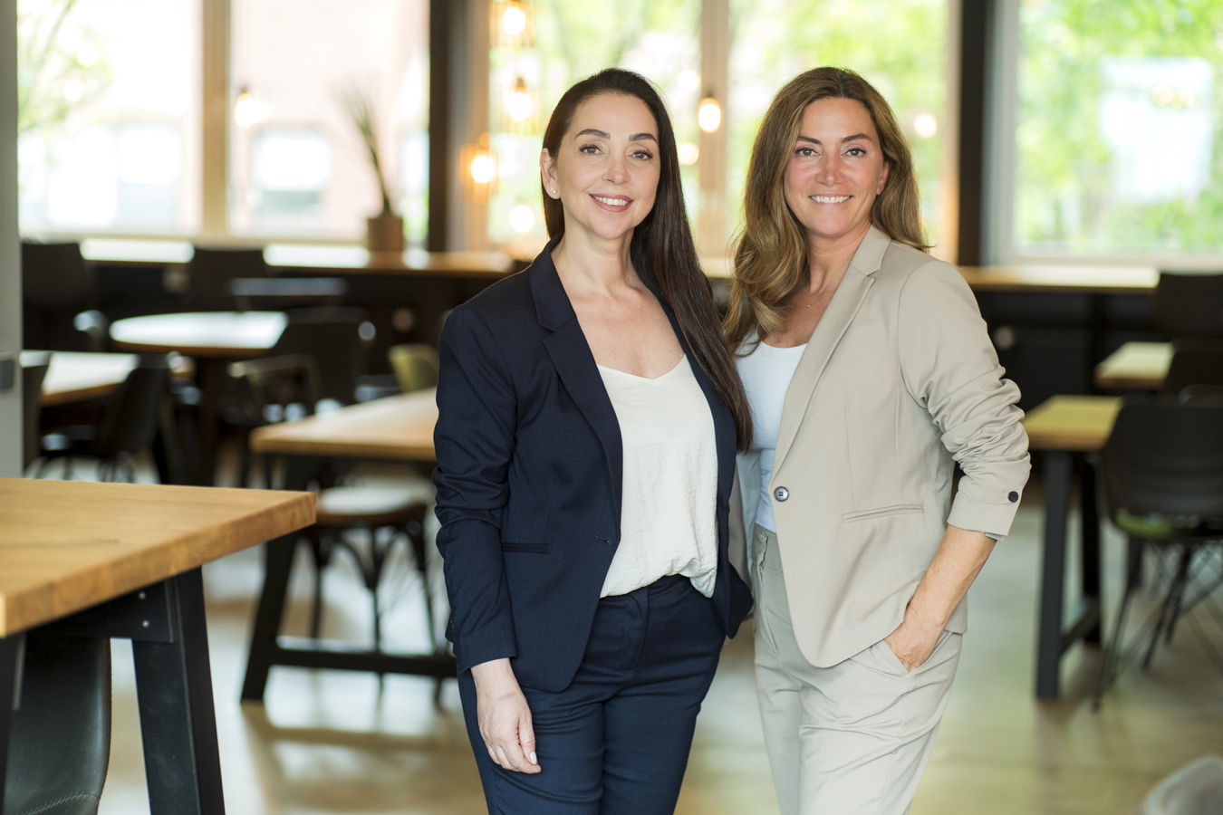 Zwei lächelnde Frauen in Businesskleidung stehen in einem hellen Café oder Bürobereich
