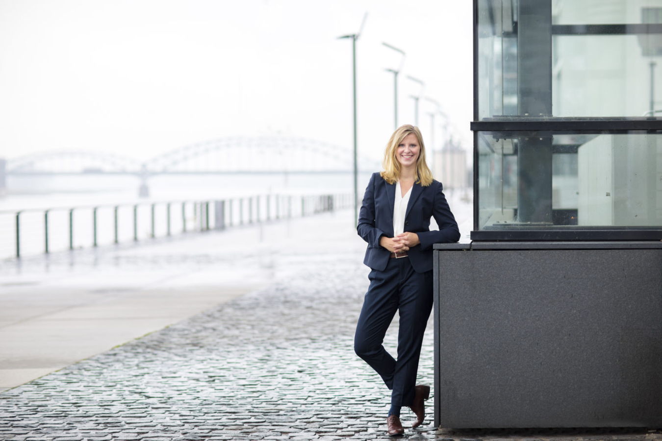 Eine Frau im Anzug lehnt an einem Glasgebäude an der Uferpromenade