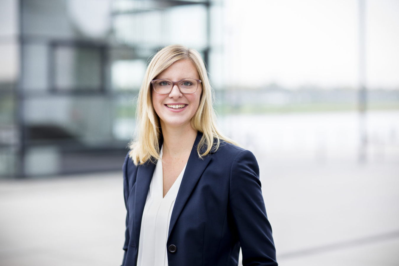 Porträt einer lächelnden Frau mit Brille im Business-Outfit vor modernem Gebäude