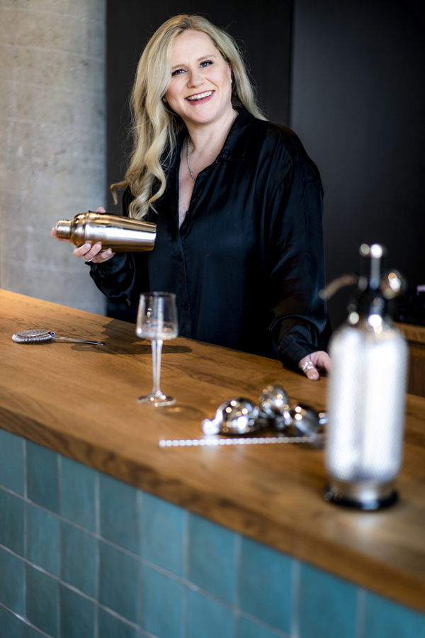 Eine lächelnde Frau hinter einer Bar hält einen Cocktailshaker