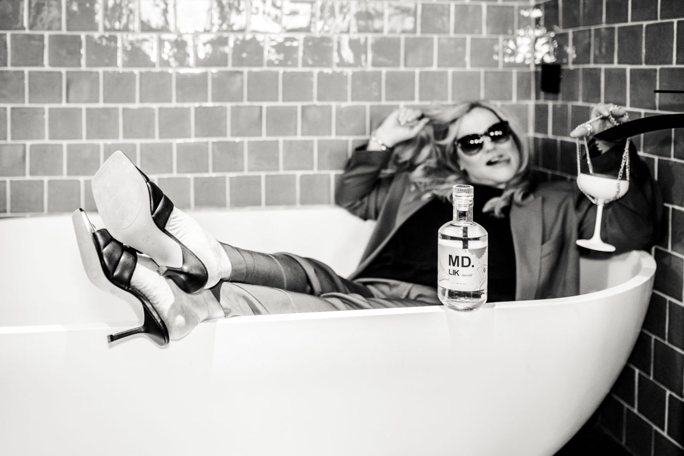 Frau mit Sonnenbrille liegt in einer Badewanne, hält ein Glas und eine Flasche MD Likör