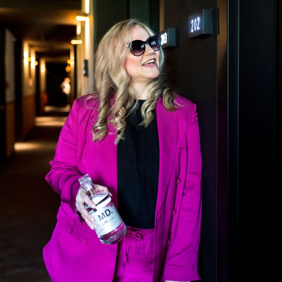 Frau in pinkem Anzug mit Sonnenbrille hält eine Flasche MD und steht in einem Hotelflur