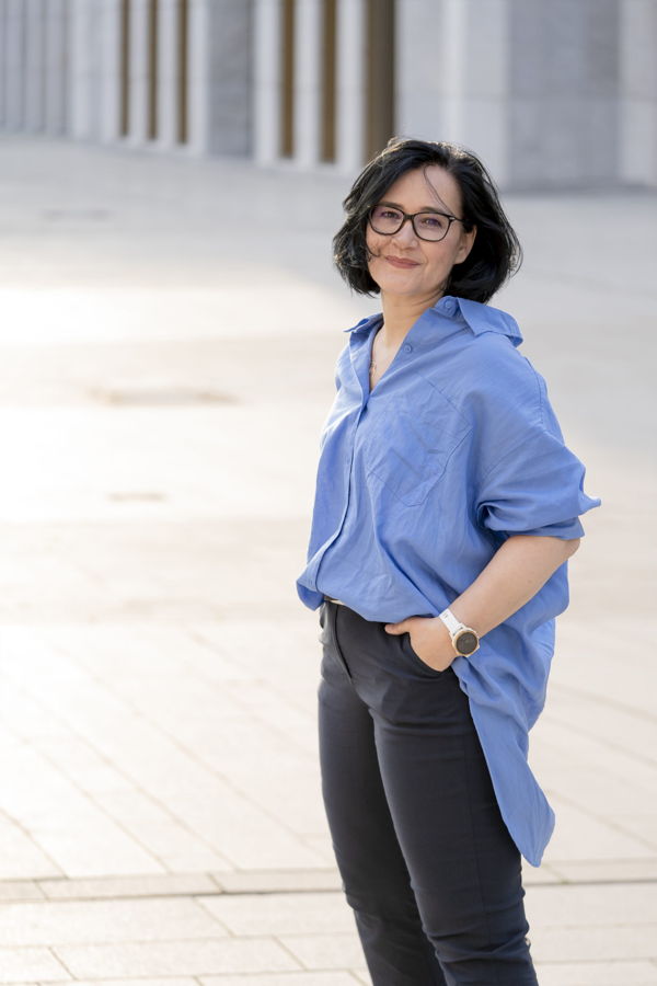 Frau mit Brille in blauem Hemd und schwarzer Hose steht im Freien