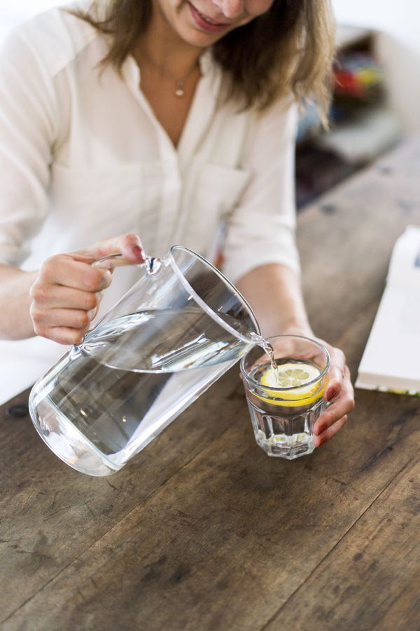 Eine Frau gießt Wasser aus einer Glaskanne in ein Glas mit Zitronenscheibe