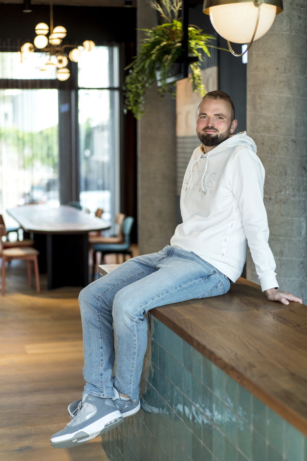 Mann in weißem Hoodie und Jeans sitzt auf einer Theke in einem Café