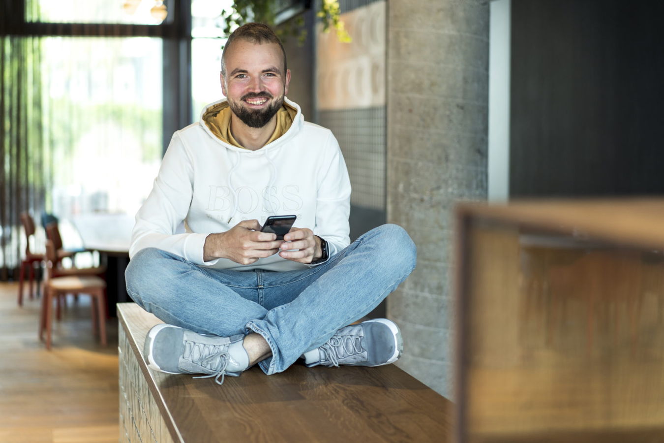 Mann sitzt lächelnd im Schneidersitz auf einer Bank und hält ein Smartphone in der Hand