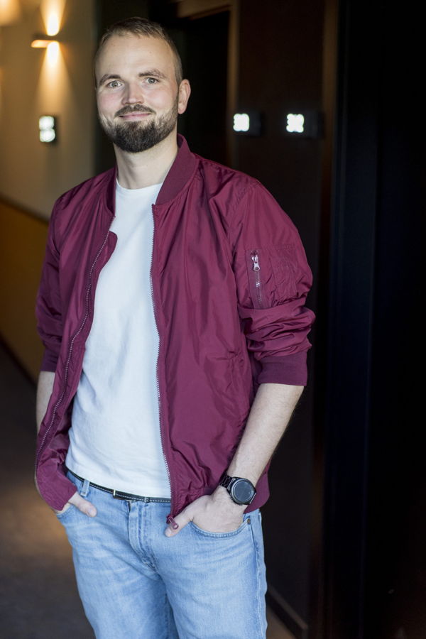 Mann mit bordeauxfarbener Jacke, weißem T‑Shirt und Jeans steht in einem Flur