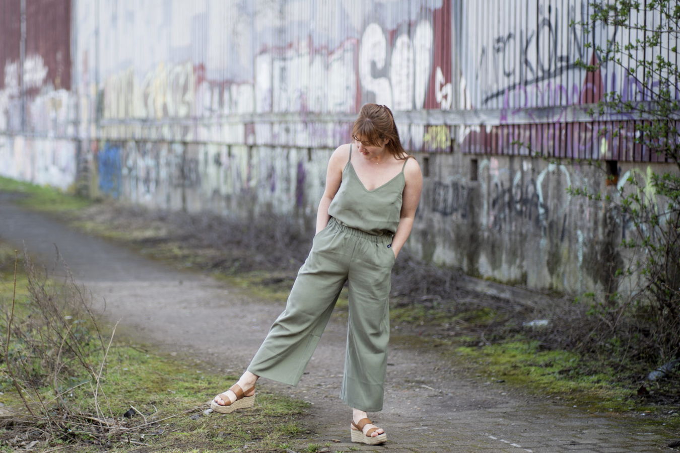 Eine Frau in einem olivgrünen Jumpsuit steht auf einem Weg vor einer Graffiti-Wand