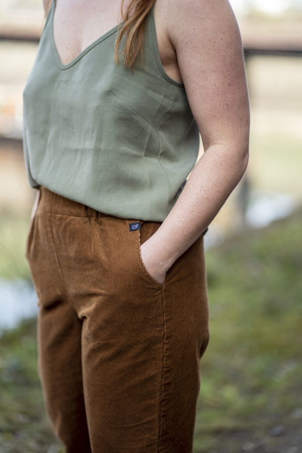 Frau in grüner ärmelloser Bluse und brauner Cordhose mit Händen in den Taschen