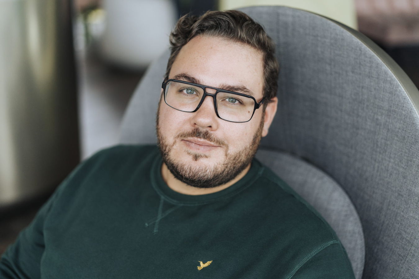 Porträt eines Mannes mit Brille in einem grünen Pullover, der in einem grauen Sessel sitzt